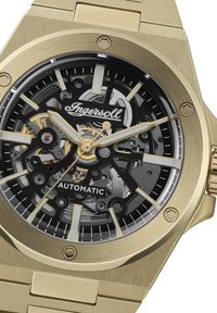Goudkleurig automatisch horloge met een zwart skeletdial, zilveren wijzers en een roestvrijstalen armband. Opmerkelijke logodetail op de wijzerplaat.