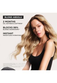 Frau mit langen, welligen blonden Haaren, die ein schwarzes Tanktop trägt. Begleitender Text hebt die Vorteile des Haarpflegeprodukts für 2 Monate Weichheit und Feuchtigkeit hervor.