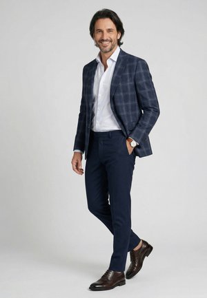 Man glimlachend, draagt een marineblauwe geruite blazer, wit overhemd, donkere broek, bruine nette schoenen en een polshorloge, staand tegen een effen lichte achtergrond.