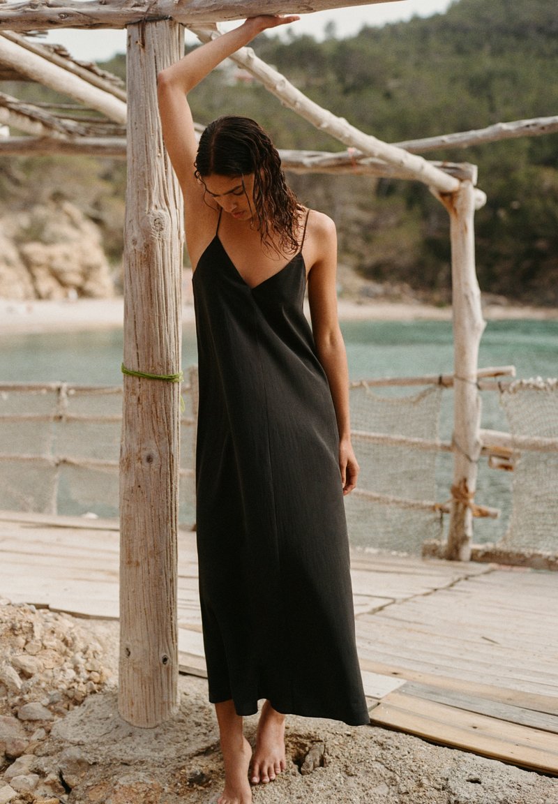 Woman in a black sleeveless maxi dress stands barefoot on sandy ground, leaning with one arm on a wooden post by a coastal dock.