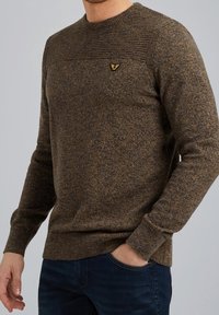 Homme portant un pull marron texturé en tricot avec empiècement côtelé et petit logo oiseau doré brodé sur la poitrine, assorti à un jean bleu foncé.