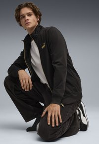 Giacca nera con zip, colletto alto e logo dorato, abbinata a jeans scuri e sneakers nere. La texture è liscia, con polsini a coste.