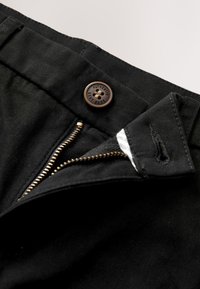 Gros plan sur la ceinture d'un pantalon noir montrant une fermeture éclair en métal partiellement ouverte et un bouton marron avec le texte "Authentic Next Dude".