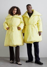 Two individuals wearing oversized, yellow puffer coats with bold, blue vertical stripes. The coats have wide collars and adjustable belts.