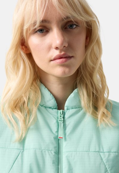 Ung kvinde med blondt hår iført en lyse mintgrøn quiltet jakke lynet op til halsen foran en ensfarvet baggrund.