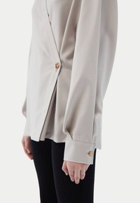 Beige långärmad skjorta med omlott-design, med en enda knapp i trä och en slät tygstruktur. Bärs med svarta ribbade leggings.