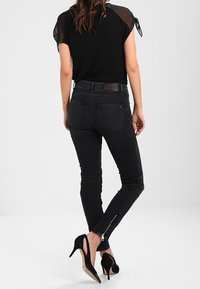Jeans skinny de mezclilla negra con cintura alta y detalles de cremallera en los tobillos. Combinados con una blusa negra con mangas transparentes.