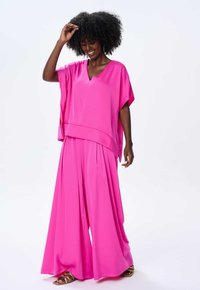 Różowy dwuczęściowy strój składający się z luźnej, krótko rękawowej bluzki z dekoltem w serek i szerokich spodni. Wykonany z gładkiego, lekkiego materiału.