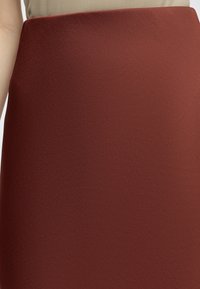 Personne portant une jupe taille haute couleur rouille et un haut beige, vue de la taille au milieu des cuisses, bras gauche partiellement visible.