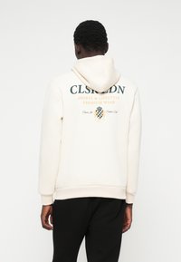 Κρέμα φούτερ με γραφικό σχέδιο στο πίσω μέρος σε πράσινο και πορτοκαλί, που διαθέτει το κείμενο "CLSRLDN SPORTS & LIFESTYLE PREMIUM WEAR". Μαλακό ύφασμα, άνετη εφαρμογή.