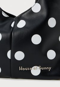 Zwarte leren tas met grote witte stippen en een zilveren "House of Sunny" logo. Glad oppervlak met een gebogen ontwerp.