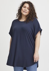 Námornícka oversize tričko vyrobené z mäkkej tkaniny, s krátkymi rolovanými rukávmi a voľným strihom s okrúhlym spodným lemom, v kombinácii s modrými džínami.