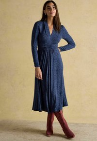 Naine pikka varrukaga, sinise mustriga midi kleidis ja põlvini punastes seemisnahast saapades, seismas ühevärvilise beeži seina vastu.