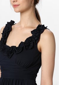Black dress with ruffled shoulder straps, fitted bodice, and gathered texture at the waist, showcasing a smooth, stretchy fabric.