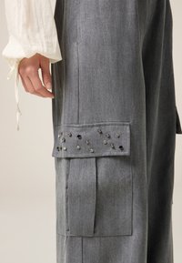 Grijze cargobroek van gestructureerde stof, met een klepzak met decoratieve studs. Rechte pijpen met geplooide details.