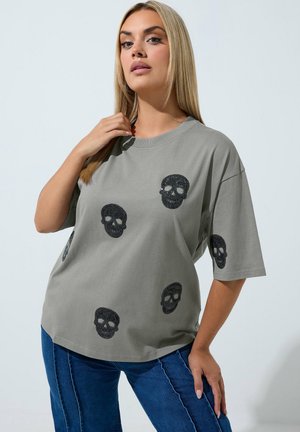 Femme aux longs cheveux blonds porte un t-shirt gris oversize avec des motifs de crânes noirs et un jean bleu, posant devant un fond clair uni.