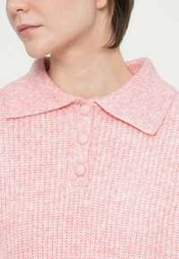 Hellrosa Strickpullover mit Kragen und vier strukturierten Knöpfen. Der Stoff weist ein geripptes Muster auf, das eine weiche Textur schafft.