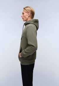 Felpa verde oliva con cappuccio regolabile, tasca frontale e polsini a costine. Indossata con pantaloni neri, il tessuto texturizzato è visibile.