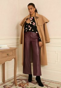 Tevegyapjú keverék kabát széles gallérral, fekete virágmintás pulóver és magas derekú burgundi bőrnadrág, fekete magassarkú bokacsizmával párosítva.