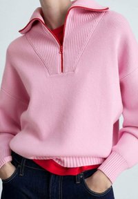 Rosa gerippter Pullover mit halbem Reißverschluss und roten Akzenten. Besitzt einen hohen Kragen und eine lockere Passform, über einem roten Shirt getragen.