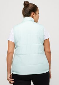 Gilet imbottito azzurro chiaro con colletto alto, design trapuntato e texture liscia. Indossato sopra una camicia bianca, presenta una silhouette aderente.