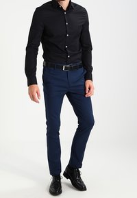 Camicia nera con bottoni, pantaloni slim-fit blu navy, scarpe eleganti in pelle nera. Caratteristiche includono una parte anteriore liscia, una cintura con accessori in argento e maniche lunghe.