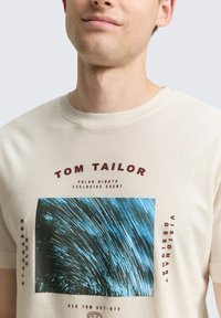 Beżowa bawełniana koszulka z niebieskim i czarnym abstrakcyjnym nadrukiem graficznym. Tekst w kolorze bordowym brzmi "TOM TAILOR" oraz "WYJĄTKOWE WYDARZENIE POLAR NIGHTS."