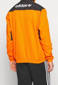 Felpa sportiva arancione con accenti neri sulle spalle, dotata di zip al collo, tasche laterali e logo Adidas bianco sul retro.
