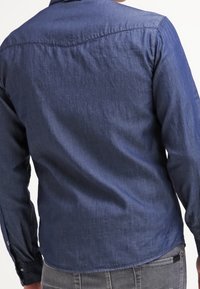 Ciemnoniebieska koszula dżinsowa z długimi rękawami, zapinanymi mankietami i subtelną fakturą; posiada wygięcie w okolicy ramion oraz prosty dół.