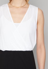 Top branco sem mangas, com um decote em V profundo, design em camadas e cintura elástica; combinado com uma saia preta. Colar em camadas dourado.
