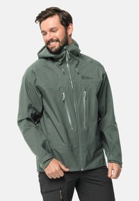 Giacca impermeabile verde realizzata in tessuto liscio, dotata di cappuccio, chiusura frontale con zip e tasche sul petto, polsini regolabili e logo sul petto.