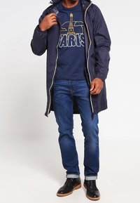 Laivastonsininen vedenpitävä takki, jossa on vetoketjullinen etuosa ja säädettävä huppu, päällä laivastonsininen grafiikkat-paita, jossa on Eiffel-torni, yhdistettynä sinisiin farkkuihin ja mustiin saappaisiin.