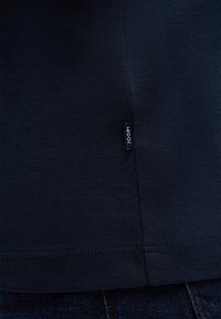 Marineblaue Baumwoll-T-Shirt mit glatter Textur, ausgestattet mit einem kleinen schwarzen Etikett mit "JOOP!" am unteren Saum. Einfaches und figurbetontes Design.