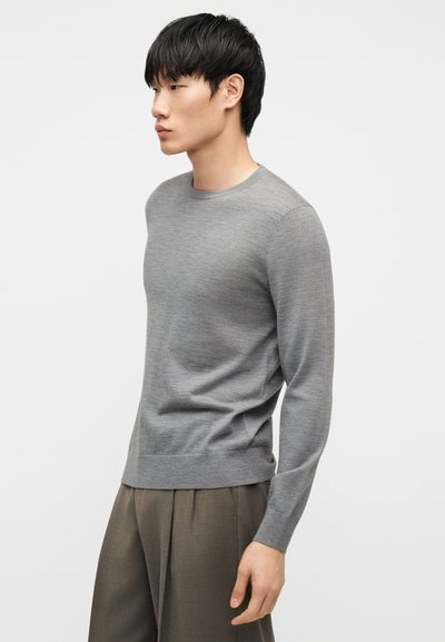 Mladý muž stojící bokem, oblečený v šedém svetru s dlouhým rukávem a volných hnědých kalhotách, na jednobarevném pozadí.