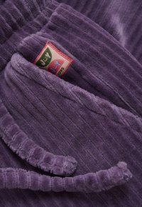 Nahaufnahme eines weichen lila Cordstoffs mit einer Tasche und einem rosa Etikett, das die "Juicy"-Marke in Grün, Pink und Blau zeigt.