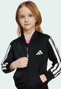 Ung tjej med ljust hår som bär en svart zip-hoodie med vita ränder och logotyp på ärmarna och bröstet, hand som justerar dragkedjan.