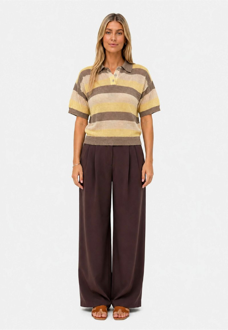 Femme debout face à l'avant, portant un polo en maille rayé à manches courtes, un pantalon large marron foncé et des sandales marron, sur un fond uni.