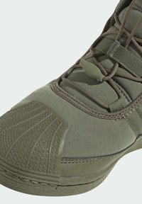 Detailní záběr na olivově zelené tenisky se zaměřením na texturovanou špičku, textilní svršek a elastické šňůrky s plastovým zámkem.