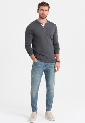 Grau langärmliges Henley-Shirt mit einer drei-Knopf-Leiste, kombiniert mit hellblauen Jeans und hellen Turnschuhen, auf einem schlichten Hintergrund stehend.