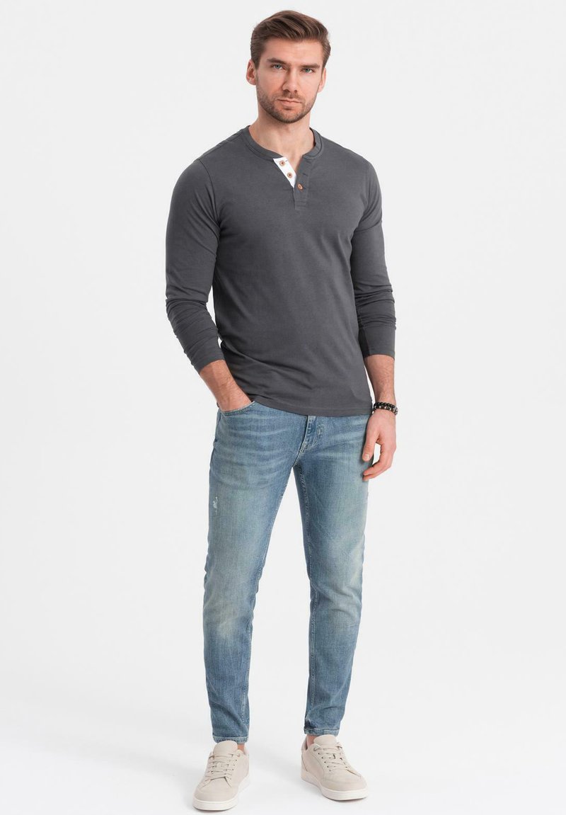 Grau langärmliges Henley-Shirt mit einer drei-Knopf-Leiste, kombiniert mit hellblauen Jeans und hellen Turnschuhen, auf einem schlichten Hintergrund stehend.
