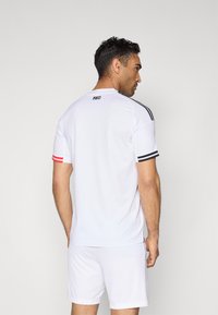 Camisola desportiva branca com detalhes em preto e vermelho nas mangas. Apresenta um decote redondo e o logótipo "1903" nas costas, confeccionada em tecido respirável.