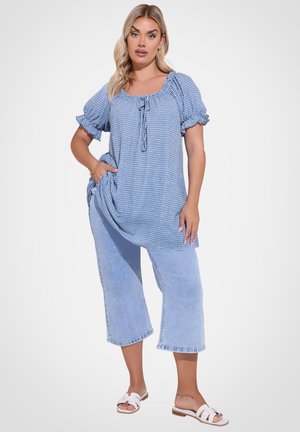 Vrouw draagt lichtblauw geruite blouse met pofmouwen, lichtblauwe cropped jeans en witte slip-on sandalen, staat met hand in de zak.