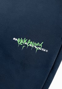 Navyblauwe katoenen sweatpants met een groene graffiti-stijl logo en witte tekstaccenten. Gladde textuur met een ontspannen pasvorm ontwerp.