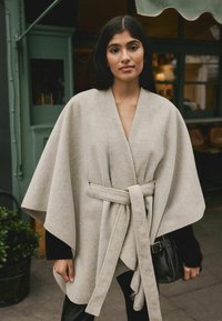 Bež mantel, mis on valmistatud pehmest kangast, laia silueti ja vööga talje kohtades, ning avatud külgedega. Kombineeritud musta kotiga.