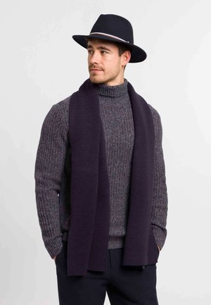 Mann trägt schwarzen Fedora, dunkelvioletten Schal und grauen, gerippten Rollkragenpullover, Hände in den Taschen, schaut seitlich vor weißem Hintergrund.