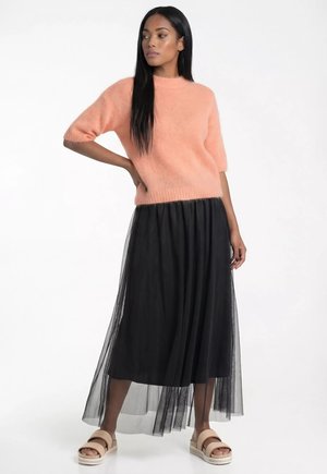 Kobieta ubrana w brzoskwiniowy, miękki sweter z krótkim rękawem, czarną, przezroczystą warstwową spódnicę midi i beżowe sandały, stojąca z jedną ręką na biodrze.