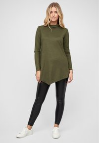 Tunique olive verte à manches longues avec un col haut, ourlet asymétrique, associée à des leggings noirs brillants et des baskets blanches.