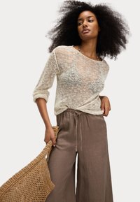 Un pull en tricot léger de couleur beige avec des manches trois-quarts, associé à un pantalon en lin marron ample. Le modèle tient un sac tissé.
