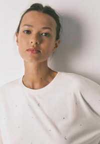 Hvid skjorte med sølvmåne- og stjernedekorationer. Glat tekstur, rund halsudskæring, afslappet pasform og korte ærmer.