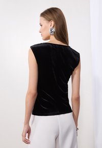 Mustmust kattega tank top, millel on vormiv disain, kõrge kaelus ja sile tekstuur, koos heledate pükstega.